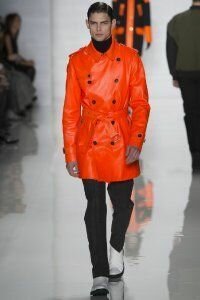 Кожаное пальто ярко-оранжевого оттенка двубортного фасона с тонким поясом от Michael Kors в сочетании с брюками чёрного цвета Michael Kors и белыми туфлями от Michael Kors.