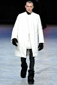 Белое пуховое пальто свободного силуэта с длиной до колен из новой коллекции Rick Owens в сочетании с джинсами чёрного цвета Rick Owens и сапогами чёрного тона с меховой отделкой от Rick Owens.