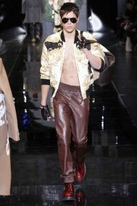 Короткое пуховое пальто для мужчин золотисто-серой расцветки от Versace в сочетании с брюками шоколадного оттенка Versace и туфлями красного тона Versace.