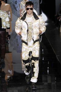 Короткое пуховое пальто на зиму шоколадно-золотистого оттенка с абстрактным рисунком, декорированное капюшоном, из новой коллекции Versace в сочетании с футболкой и брюками в тон пальто Versace и туфлями чёрного цвета от Versace.