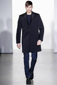 Мужское зимнее пальто чёрно-синего оттенка прямого силуэта с фактурным рисунком от Calvin Klein в сочетании с брюками тёмно-синего тона Calvin Klein и чёрными кроссовками Calvin Klein.