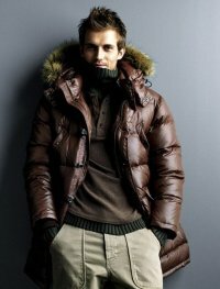 Пуховое мужское пальто на зиму шоколадного оттенка с капюшоном в сочетании с брюками светло-серого тона и свитером серо-коричневой расцветки.