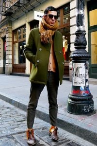 Короткое мужское пальто на зиму двубортного фасона травянисто-зелёного оттенка в сочетании с шарфом рыжего цвета, свитером бежевого тона, тёмно-серыми джинсами и туфлями рыже–коричневой расцветки.